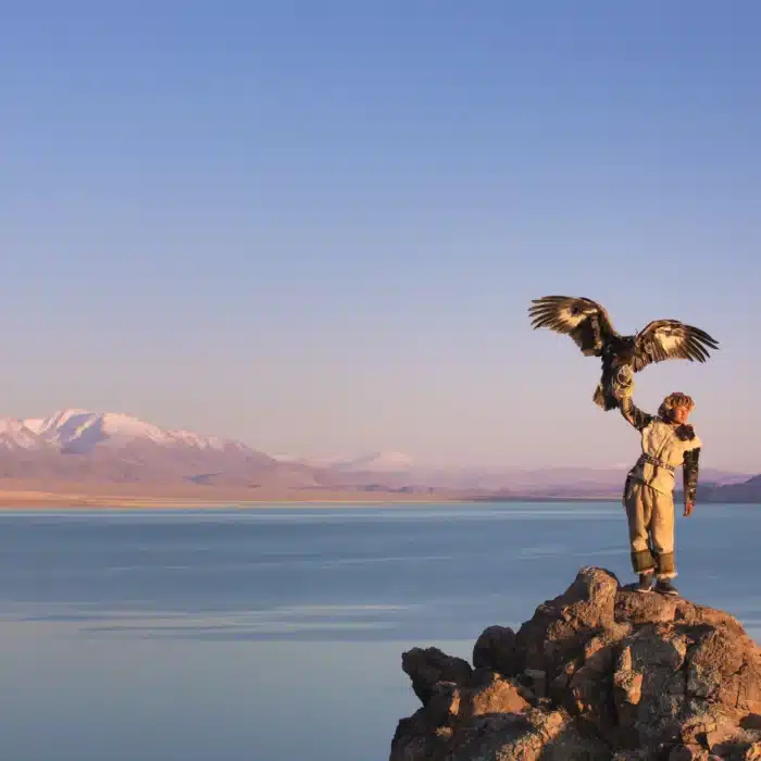 Traditional kazakh eagle hunter with his golden eagle in front of snow capped mountains at a lake shore. Ulgii, Western Mongolia.